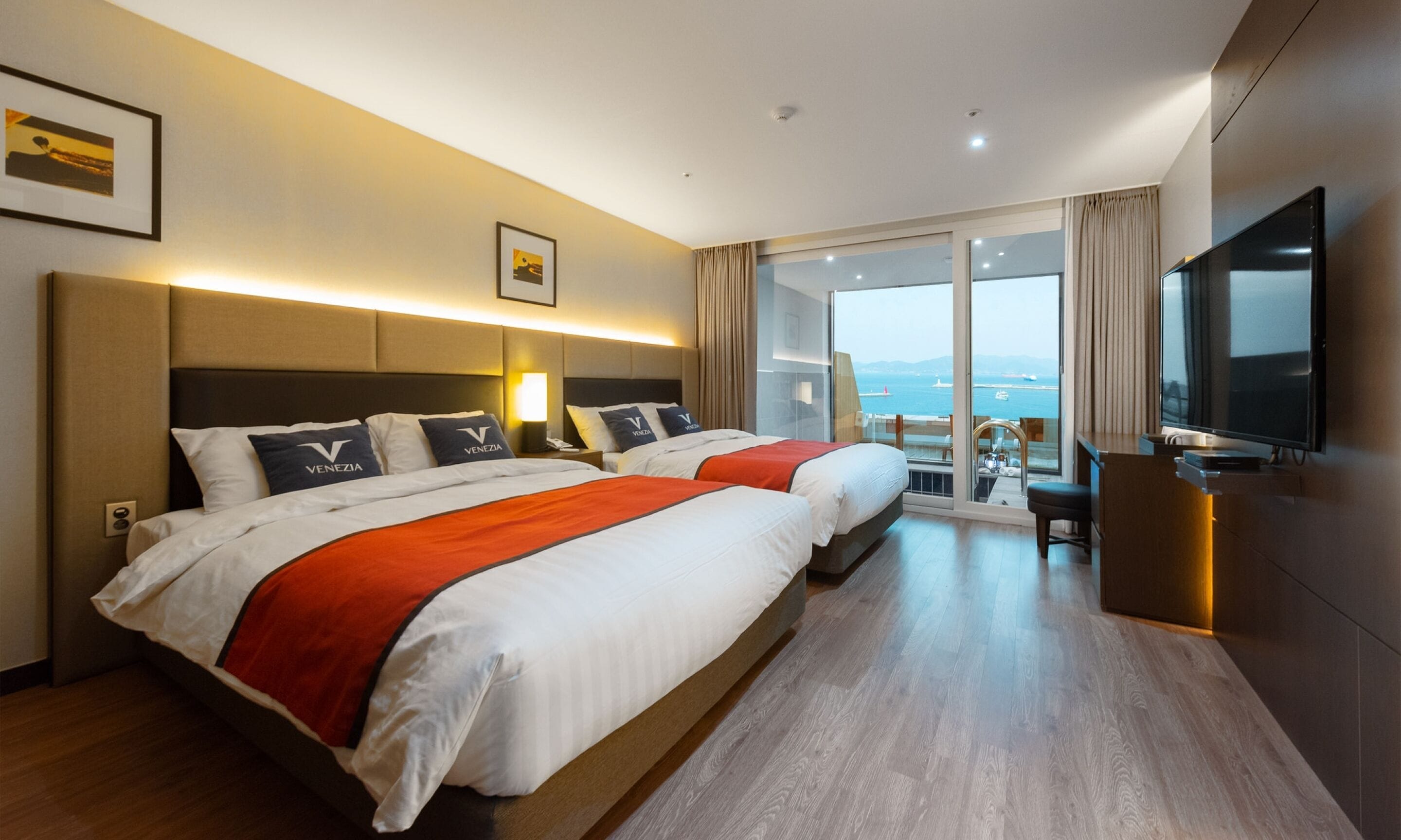 A hotel room with two beds, a TV, and a balcony with a view of the sea.