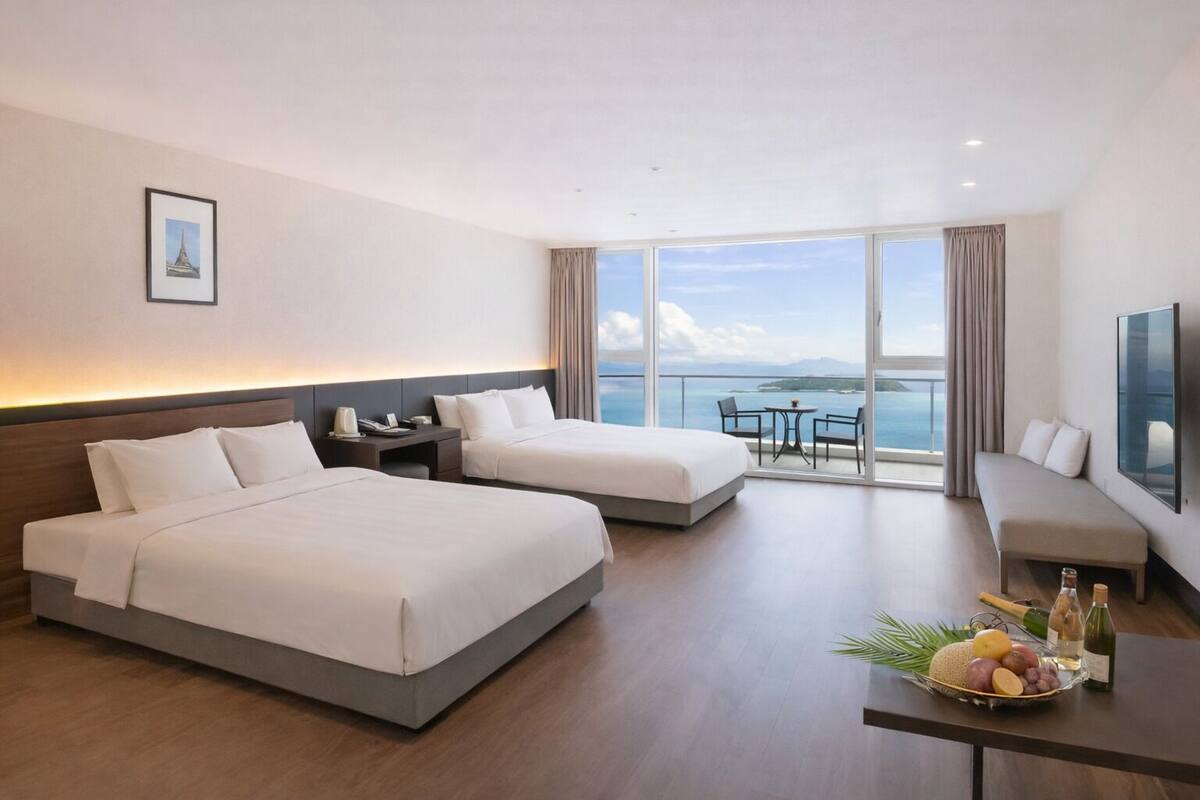 Ein Hotelzimmer mit zwei Betten, einem Balkon mit Tisch und Stühlen sowie einem großen Fenster mit Blick auf das Meer.