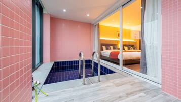 Un baño moderno con bañera, zona de ducha y puerta de vidrio que da a un dormitorio con cama y mesita de noche.