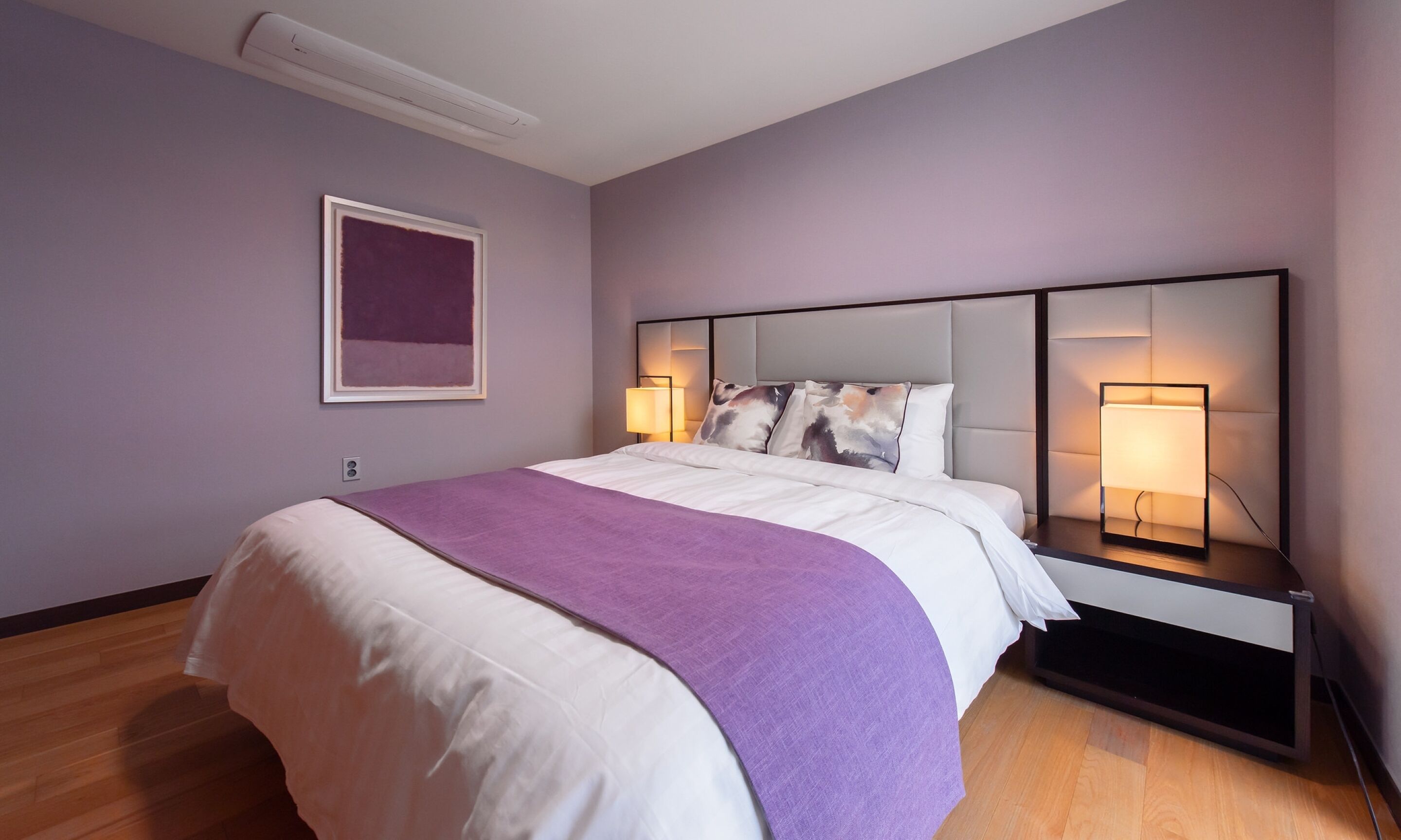 A modern hotel room with a large bed, a headboard with integrated lighting, a bedside table with a lamp, and a framed artwork on the wall.