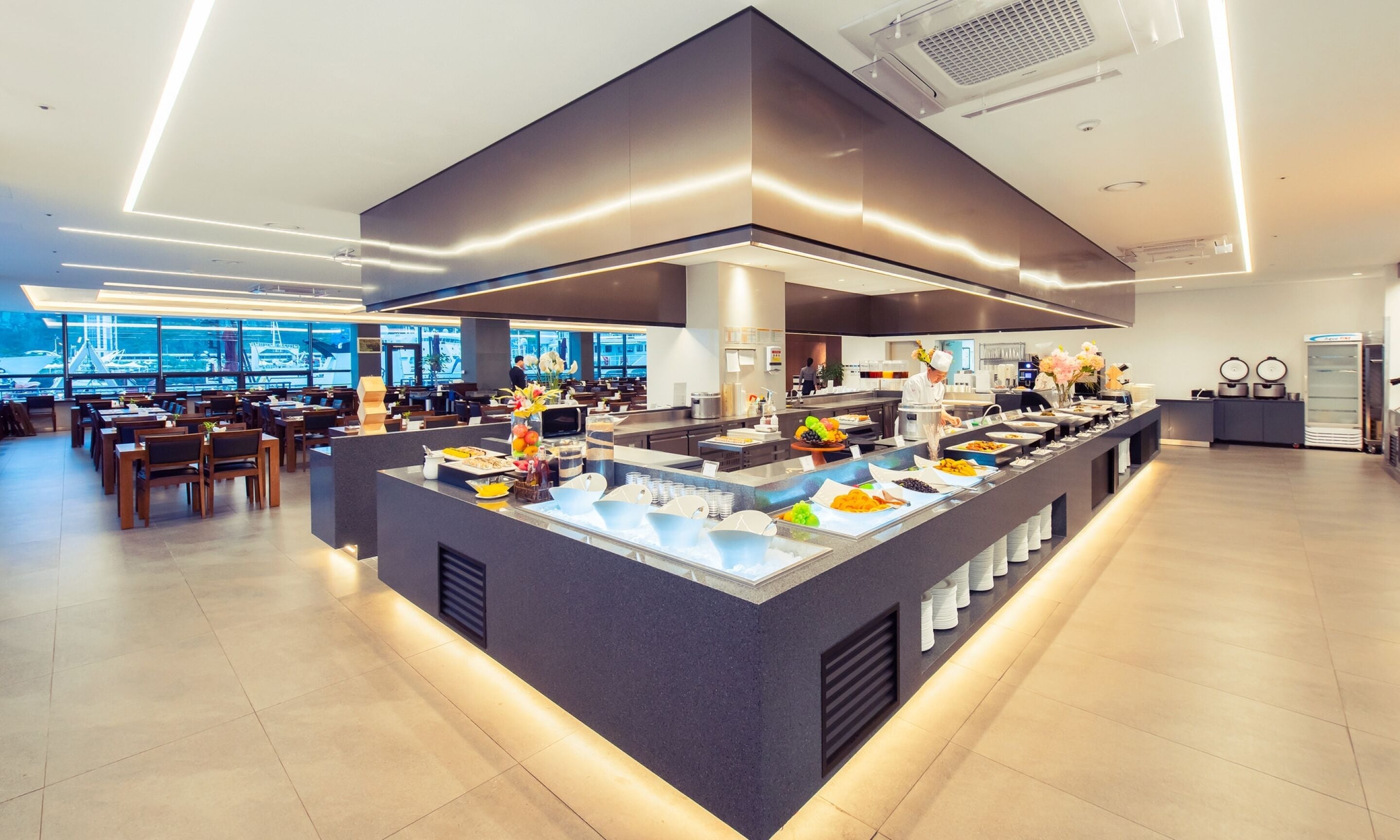 Un bar à petit-déjeuner moderne proposant une variété de plats, un chef cuisinier en train de préparer les aliments, et une vue sur l’aéroport en arrière-plan.
