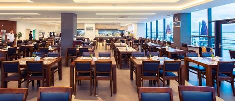 Un restaurante espacioso con varias mesas y sillas, grandes ventanales que ofrecen vistas al mar, y una decoración interior moderna.