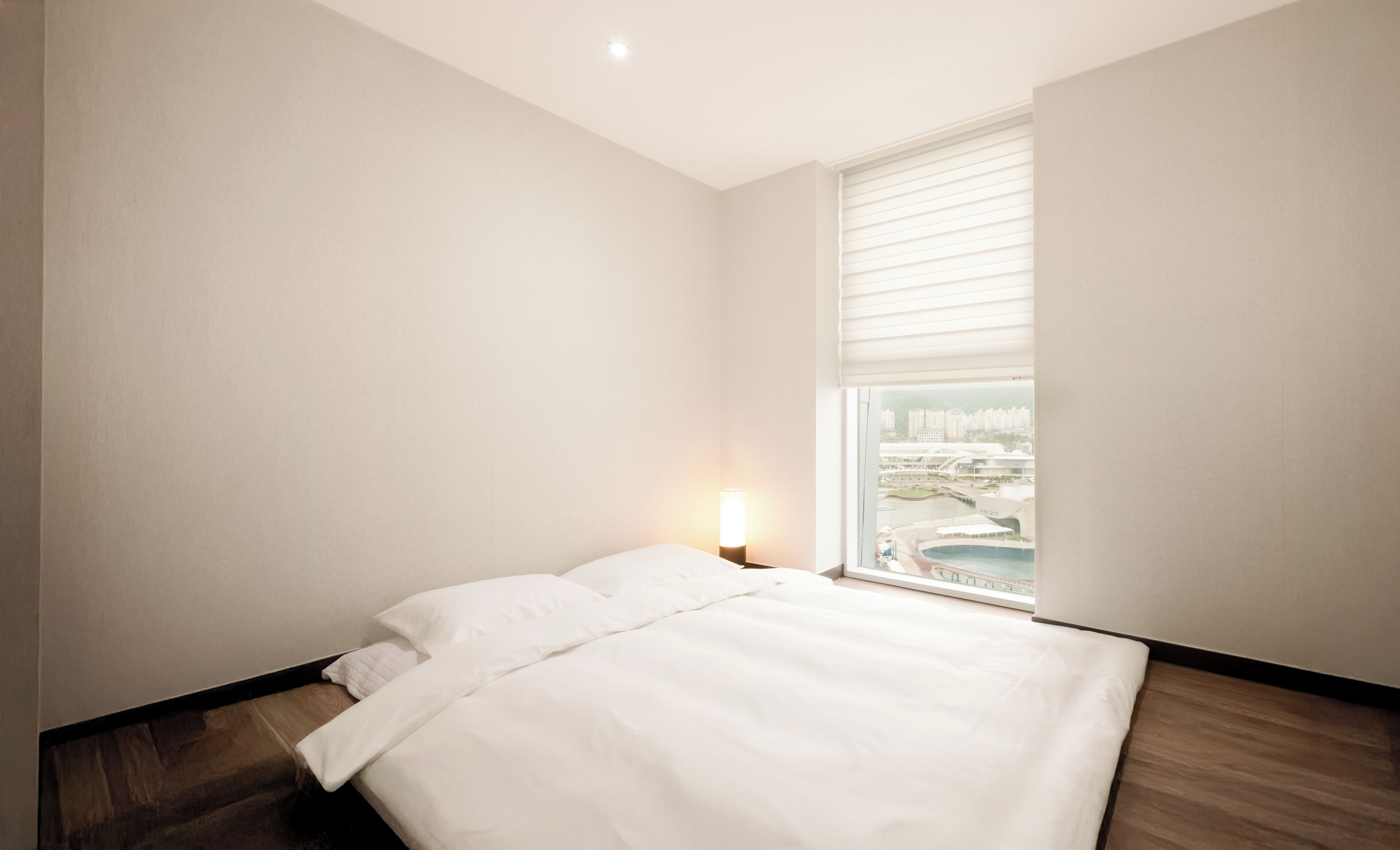 A hotel room with a bed, a nightstand, a lamp, and a window with a city view.
