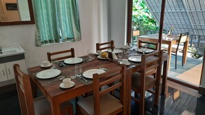 A dining area with a wooden table set for four, surrounded by chairs. A window with green curtains is visible, and a small sink is to the left.