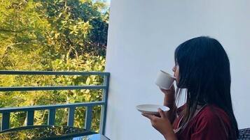 Una persona sentada en una silla de mimbre frente a una mesa redonda pequeña en un balcón con vistas a follaje verde.