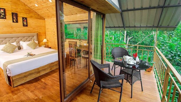 A wooden room with a bed, a dining table with chairs, and a balcony with a view of greenery.