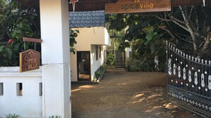 A rustic villa entrance with a tiled roof, a wooden signboard, and a metal gate.