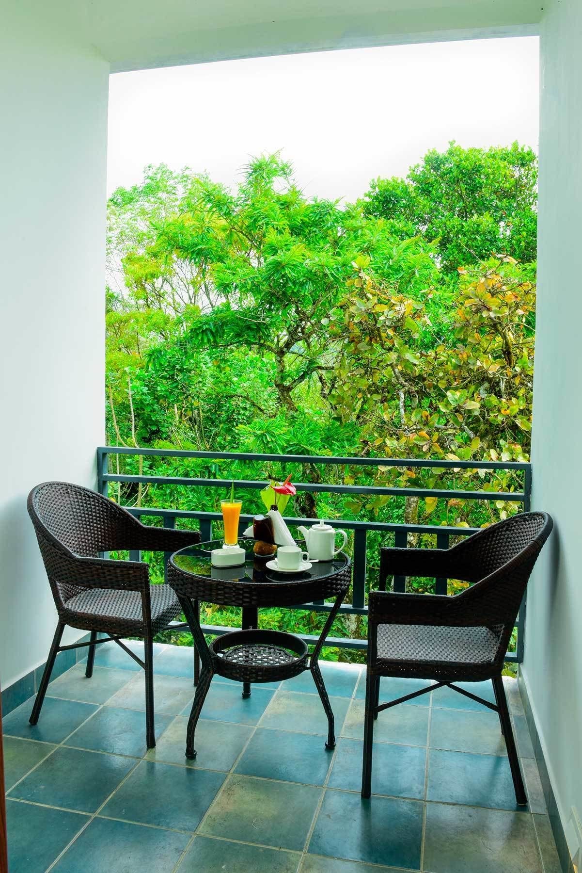 A balcony with two wicker chairs, a small round table, and a view of lush green trees.