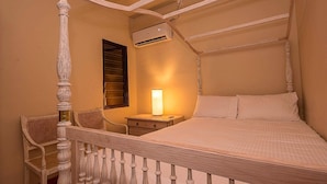 A four-poster bed with a canopy, a bedside table with a lamp, a wicker chair, and an air conditioning unit on the wall. - Arathi Appartment (Galle)