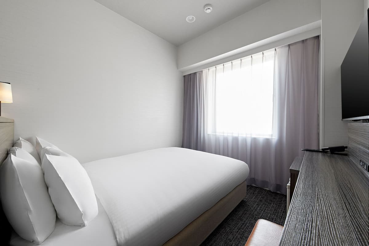 A hotel room with a bed, a desk, a lamp, and a window with curtains.