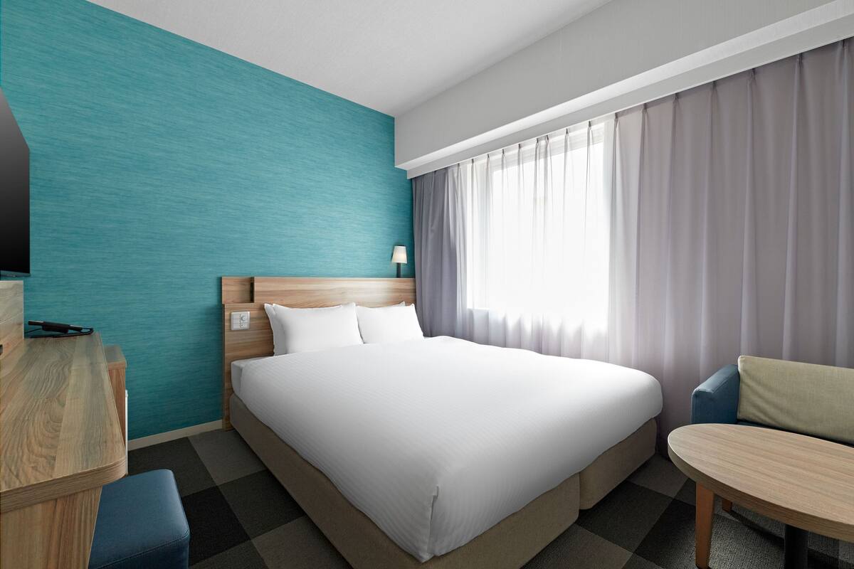 A hotel room with a large bed, a wooden desk, a small table, and a view of the outdoors through a window with curtains.