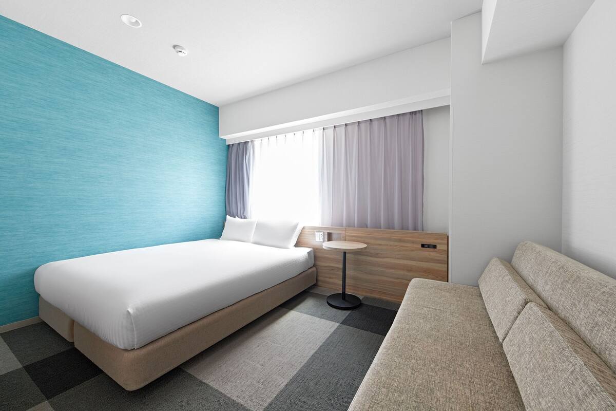 A modern hotel room with a large bed, a sofa, a small round table, and a window with curtains.