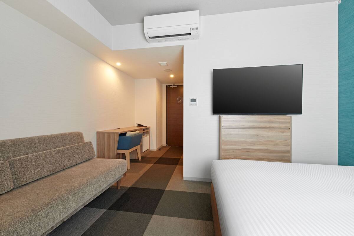 A modern hotel room with a sofa, a desk, a bed, and a mounted TV.