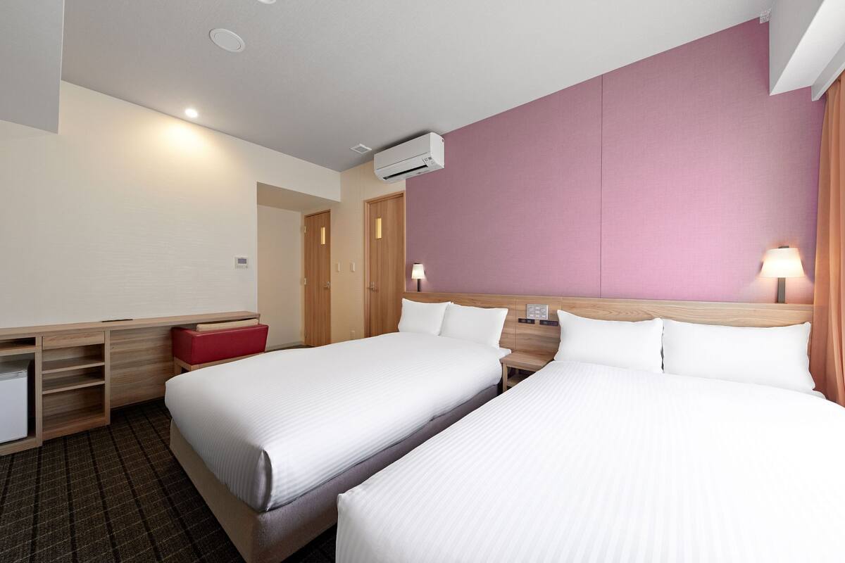 A hotel room with two beds, a desk, a red bench, and a wall-mounted air conditioner.