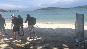 Ein Strandbild mit Menschen, die am Ufer entlanggehen, einem Segelboot im Wasser und einem Schild im Sand.
