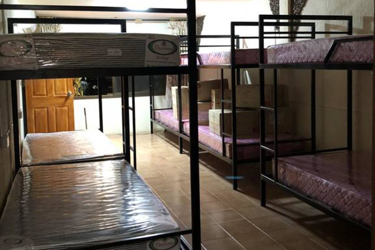 A dormitory room with bunk beds, a tiled floor, and a visible kitchen area in the background.