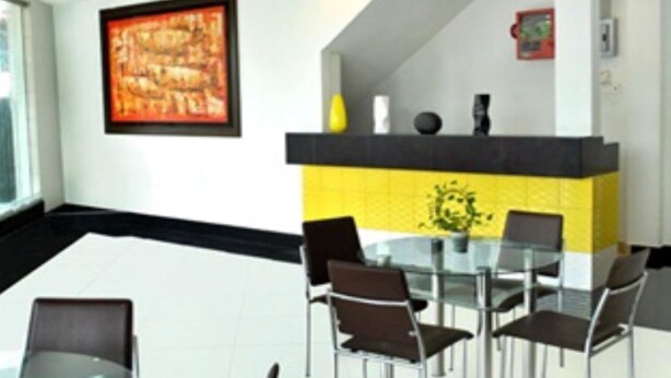 Un comedor moderno con una mesa de vidrio y sillas de metal, una pared de acento amarilla y negra, y una obra enmarcada sobre la mesa.