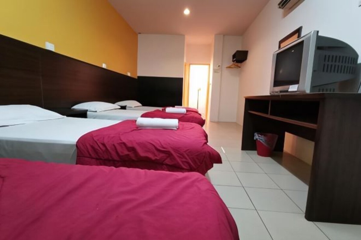 A hotel room with three beds, each with a red and white bedspread, a television on a wooden stand, and a red bucket on the floor.
