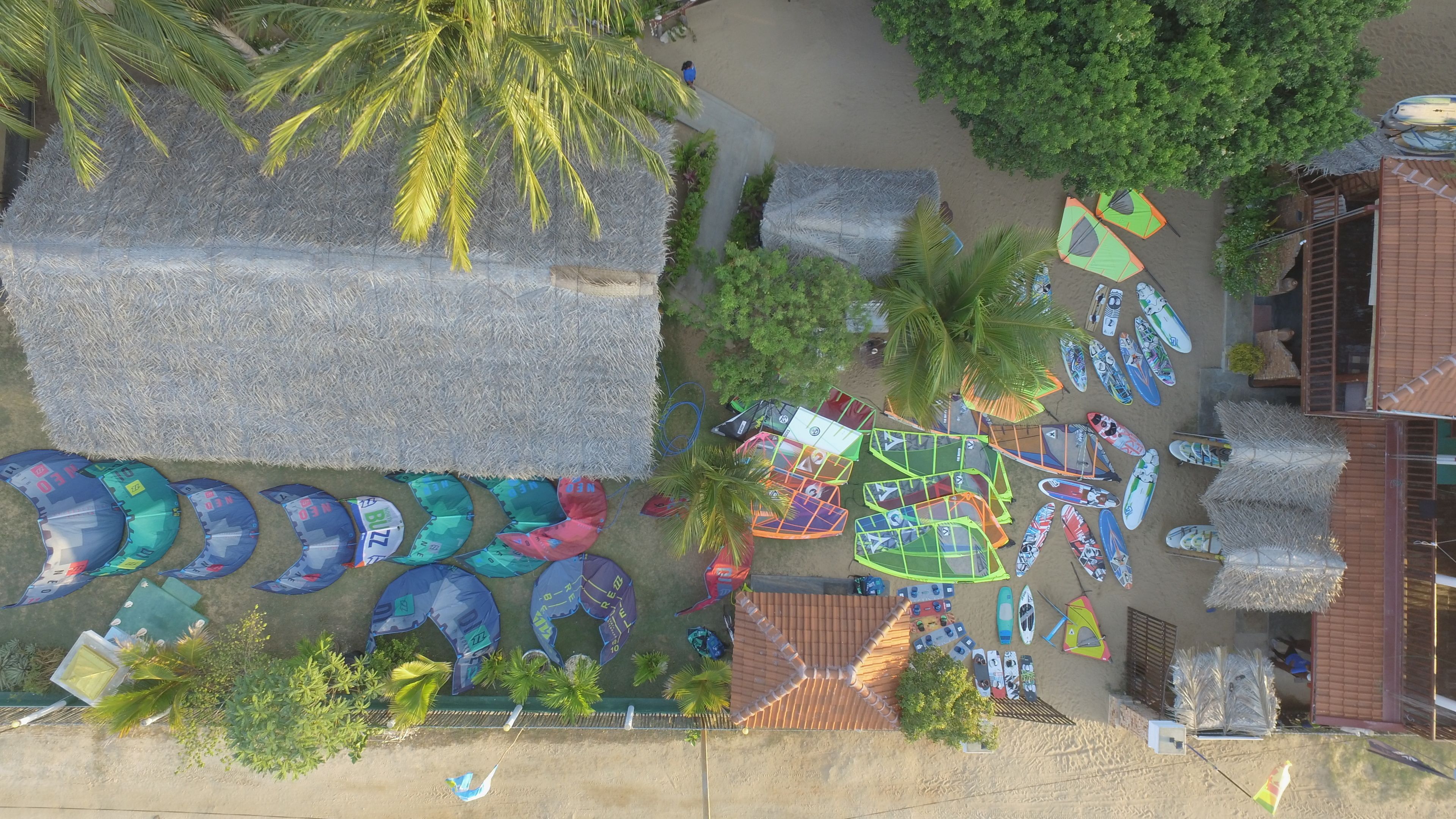 An aerial view of a beachside property with thatched-roof huts, a variety of colorful parasails, and a well-maintained garden area.