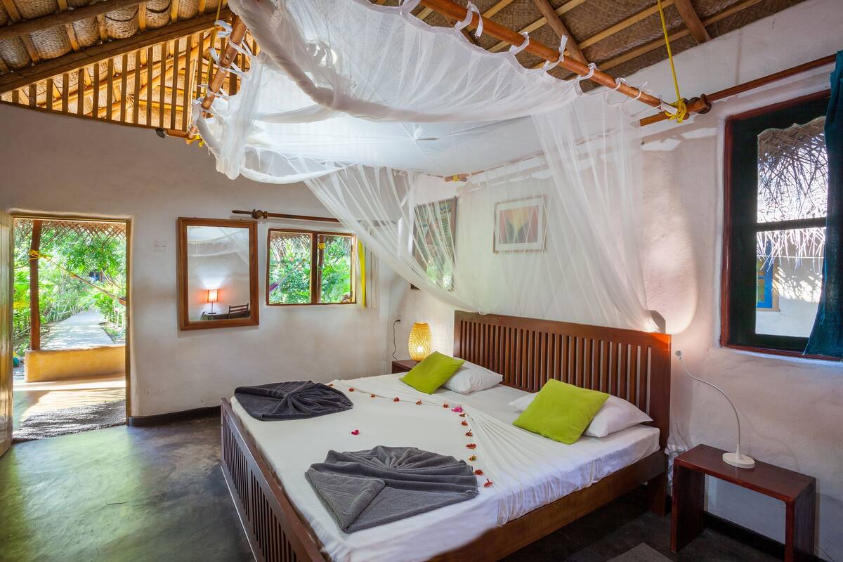 A room with a bed, bedside table, and a window with a view of greenery.