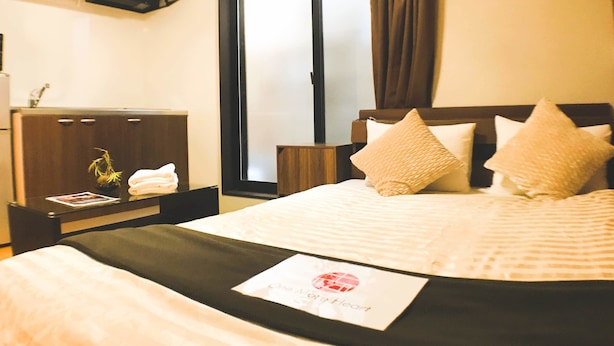 A hotel room with a bed, a bedside table with a lamp, a window with curtains, and a TV mounted on the wall. - GH One More Heart Jujo 1 (Kyoto)