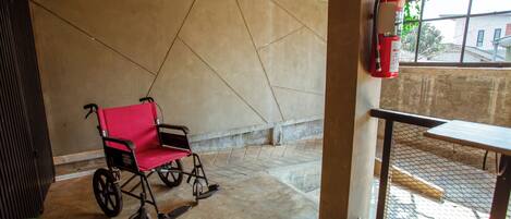 A wheelchair, a fire extinguisher, and a table in a covered outdoor area with a concrete floor and walls.