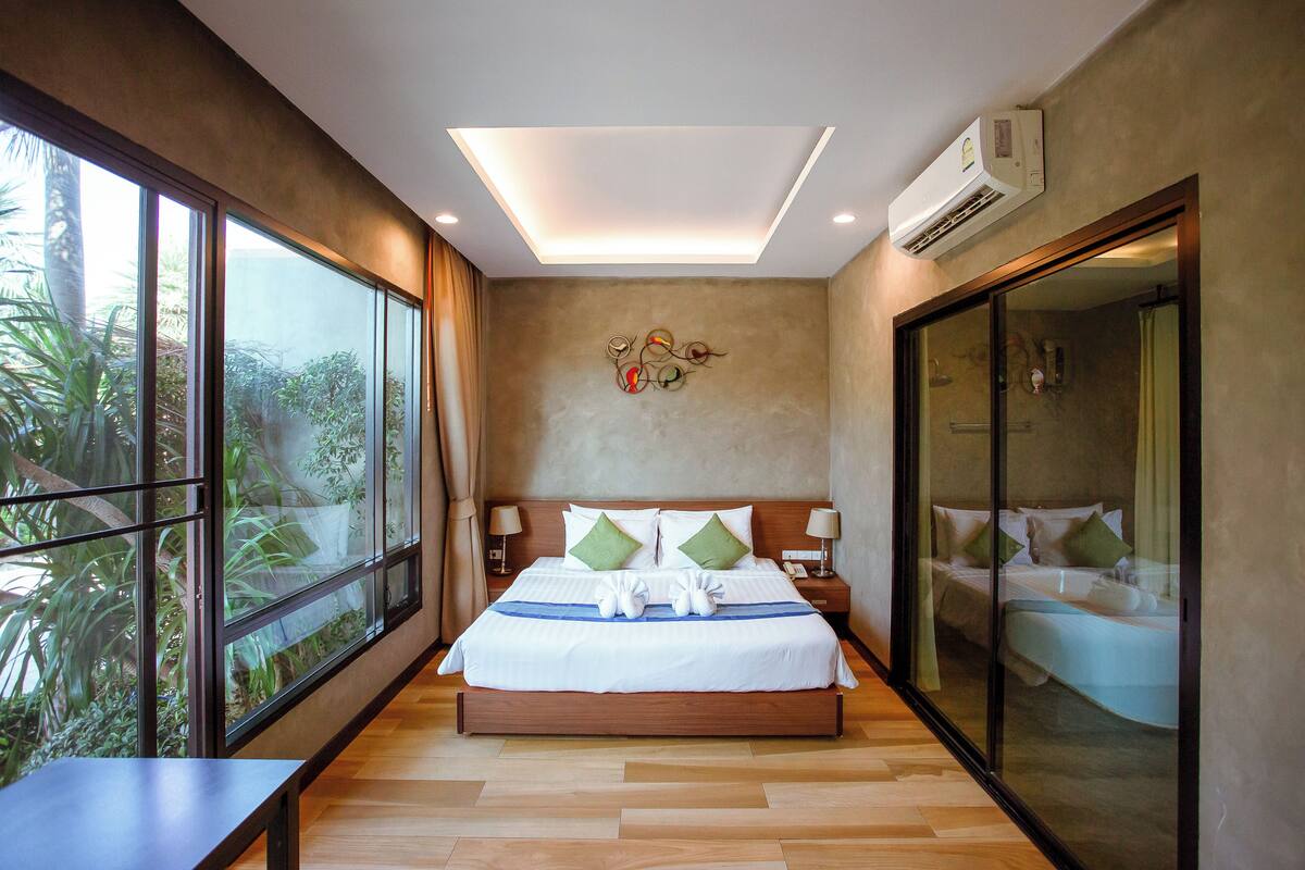 Ein modernes Hotelzimmer mit einem großen Bett, einer Deckenleuchte, einer Klimaanlage und Blick auf Grün durch große Fenster.