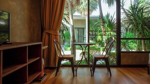 Quarto com televisão, prateleira de madeira, duas cadeiras, uma mesa pequena e uma janela grande com vista para palmeiras.