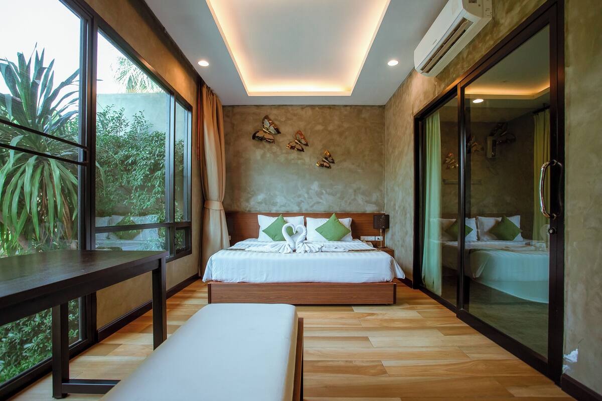 Ein modernes Hotelzimmer mit einem großen Bett, einer Sitzbank, einer Duschkabine aus Glas und Blick auf Grünflächen.
