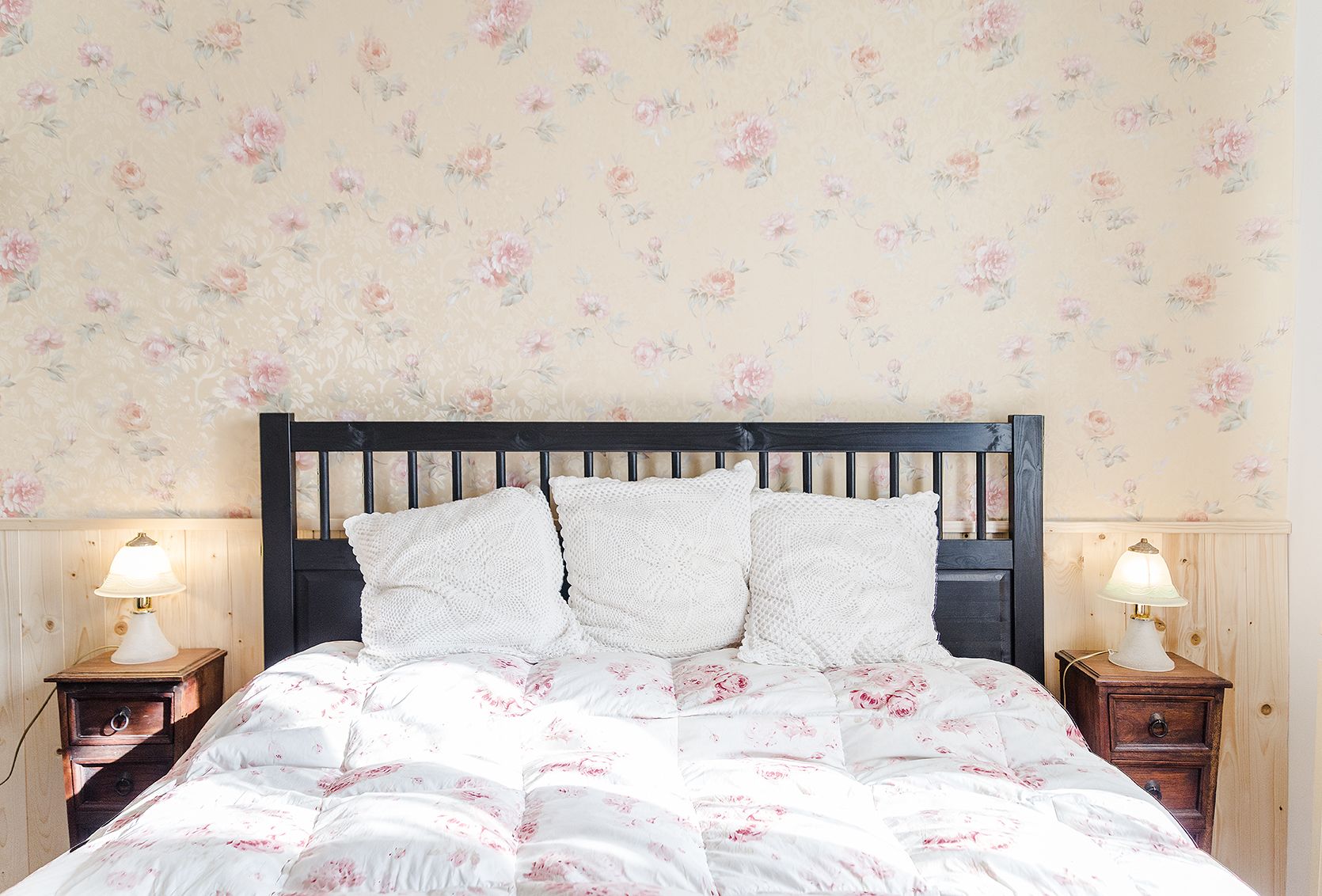 A bedroom with a floral wallpaper, a wooden headboard, white bedding, and two bedside lamps.