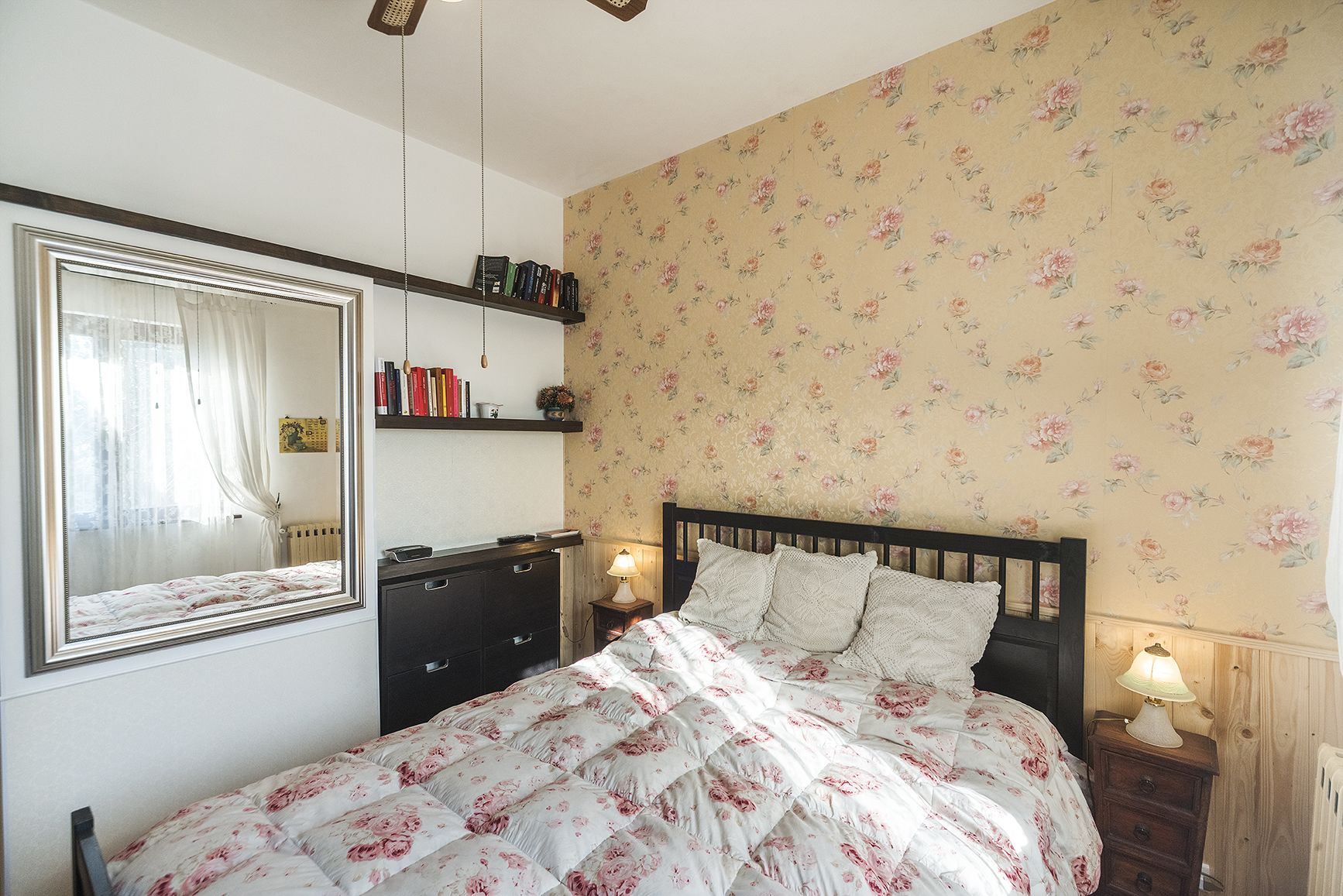 A bedroom with a bed, a dresser, a mirror, a shelf with books, and floral wallpaper.