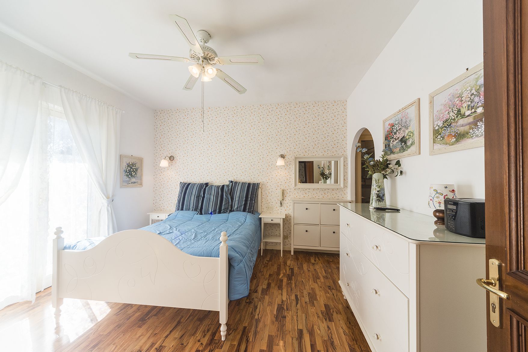 A bedroom with a bed, a desk, a dresser, a ceiling fan, and a window with curtains.