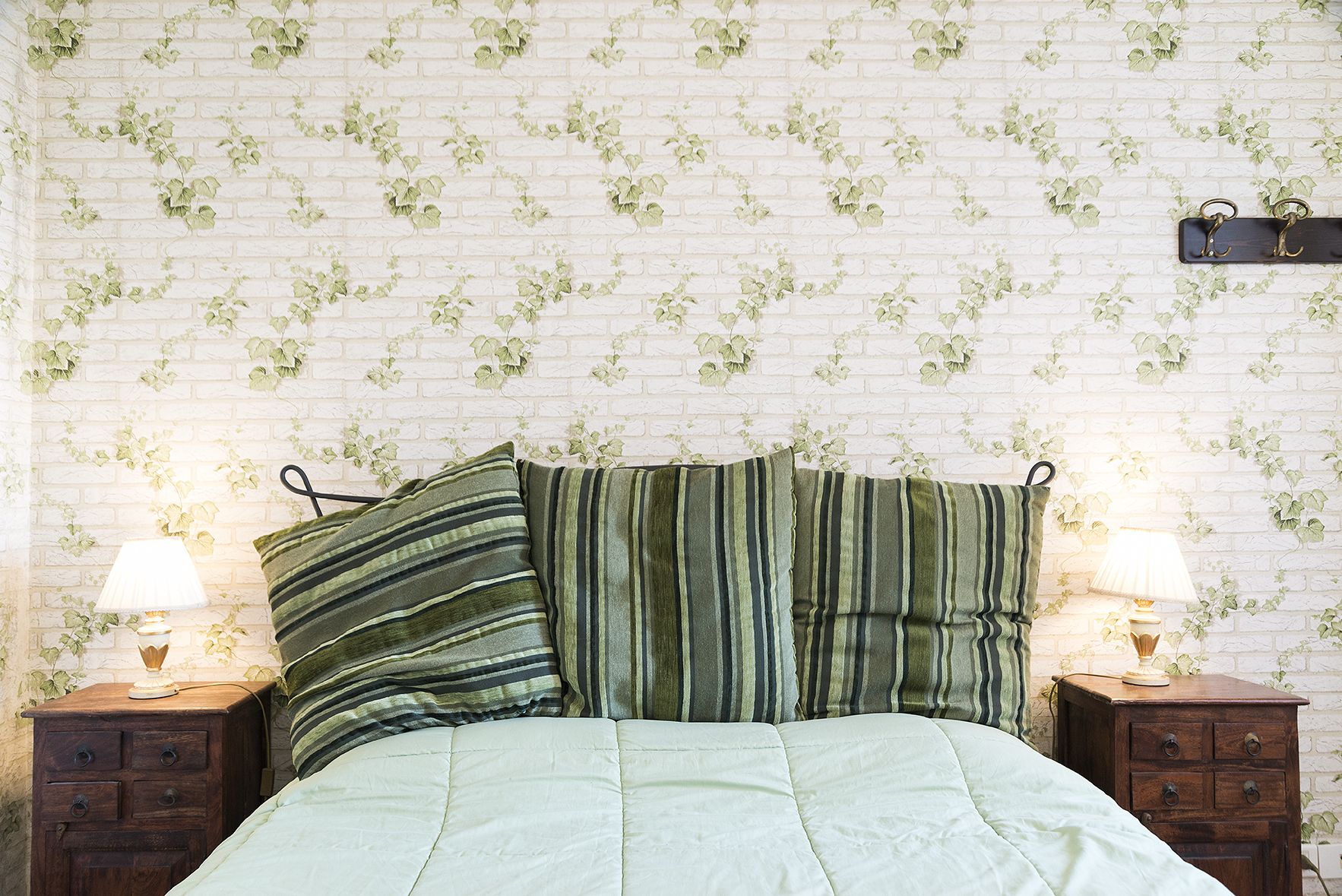A bedroom with a brick-patterned wall, a bed with striped pillows, two bedside tables with lamps, and a small wooden chest.