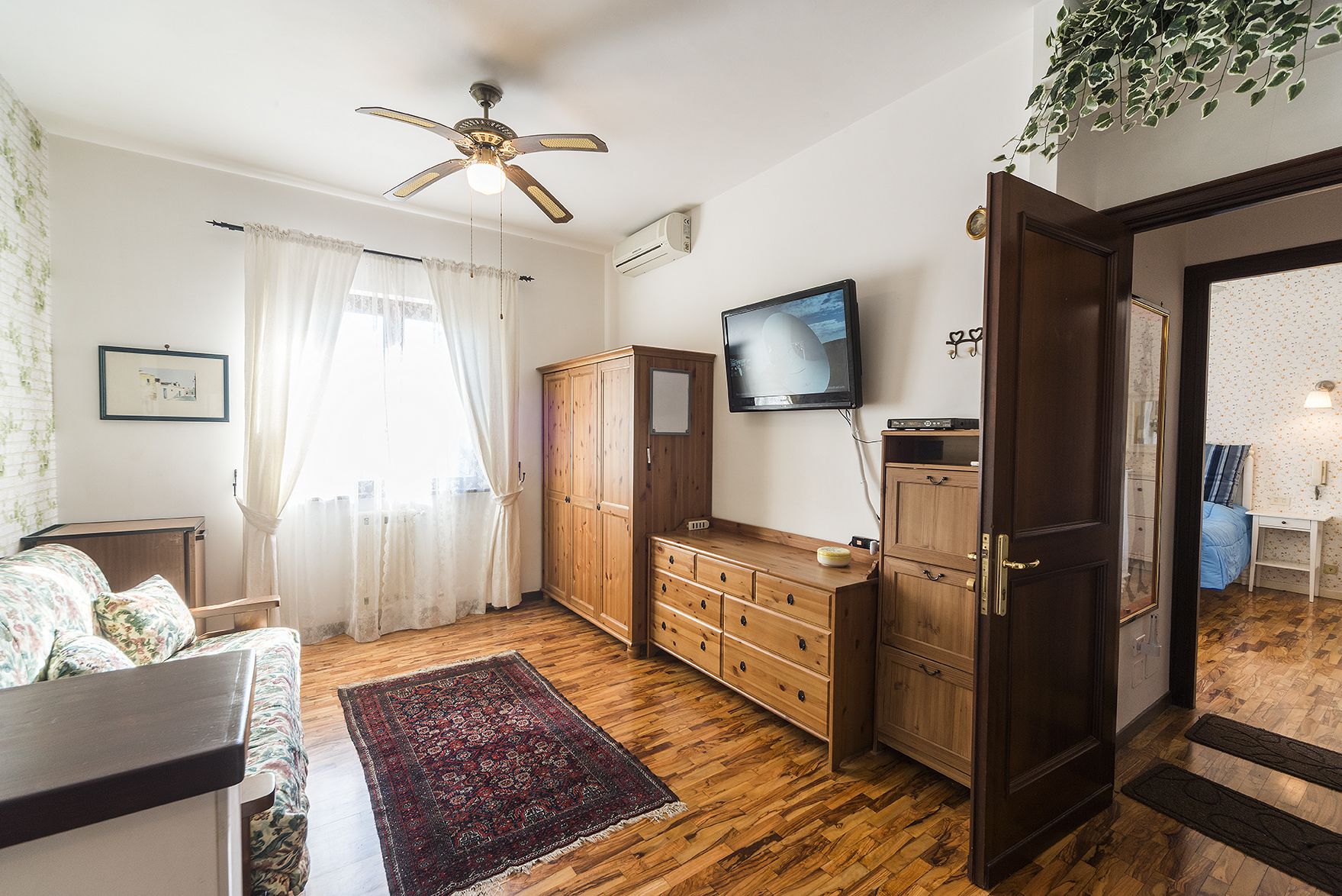 A room with a wooden floor, a ceiling fan, a TV mounted on the wall, and a door leading to another room.