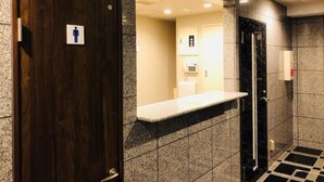 Baño de hombres con puerta de madera, espejo, pared con azulejos de mármol y puerta negra.