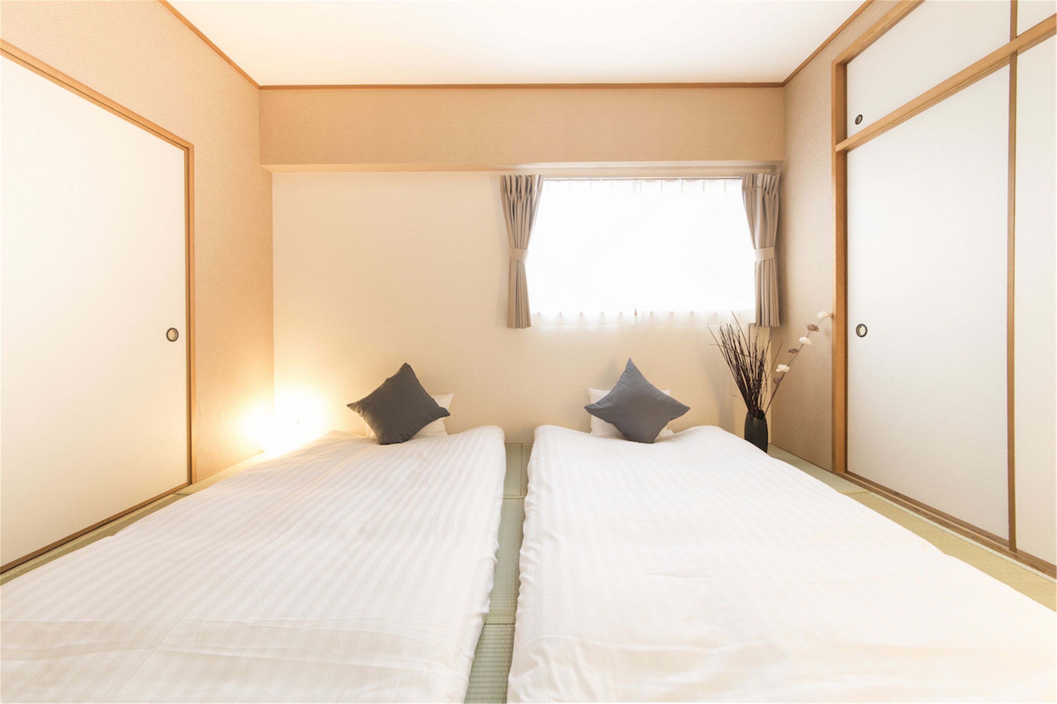 Hotellihuone, jossa on kaksi erillistä sänkyä, ikkuna, jossa on verhot, ja maljakko, jossa on kuivattuja kasveja.