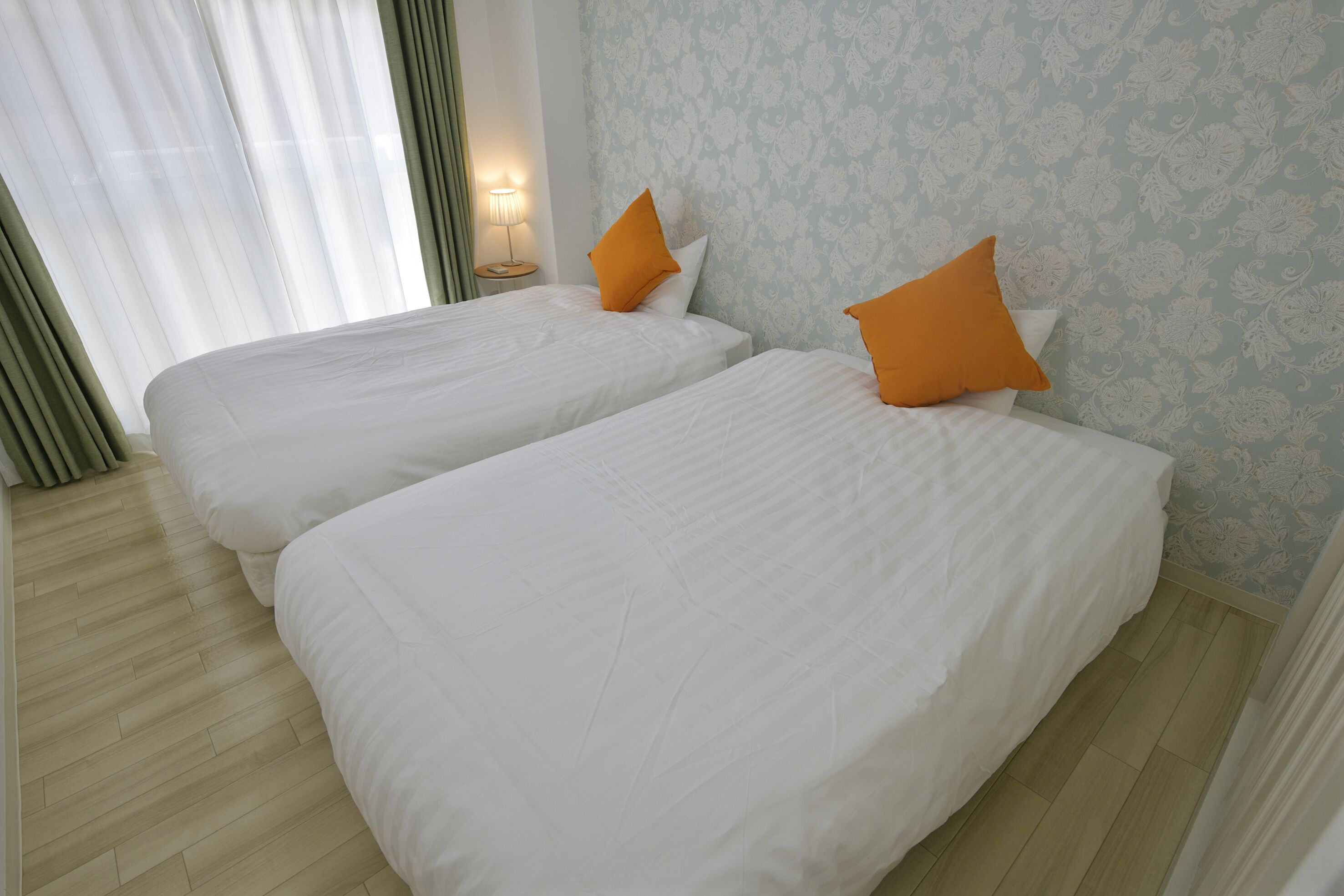 Habitación de hotel con dos camas, una mesita de noche con lámpara y un papel tapiz estampado con motivos florales.