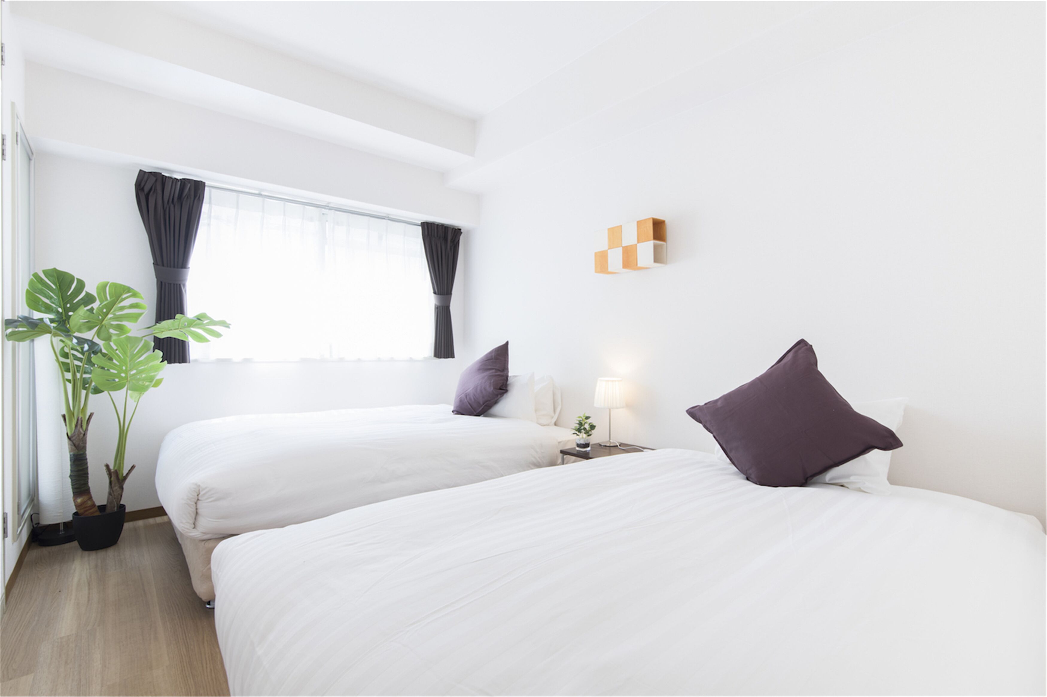 A hotel room with two beds, a potted plant, a window with curtains, a bedside table with a lamp, and a wall-mounted light fixture.