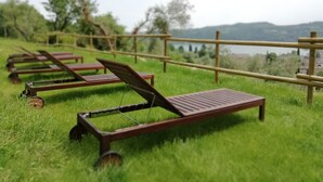 A row of wooden lounge chairs on a grassy hill with a bamboo fence, overlooking a body of water and distant hills. - Agriturismo Podere Nigriano (Salò)