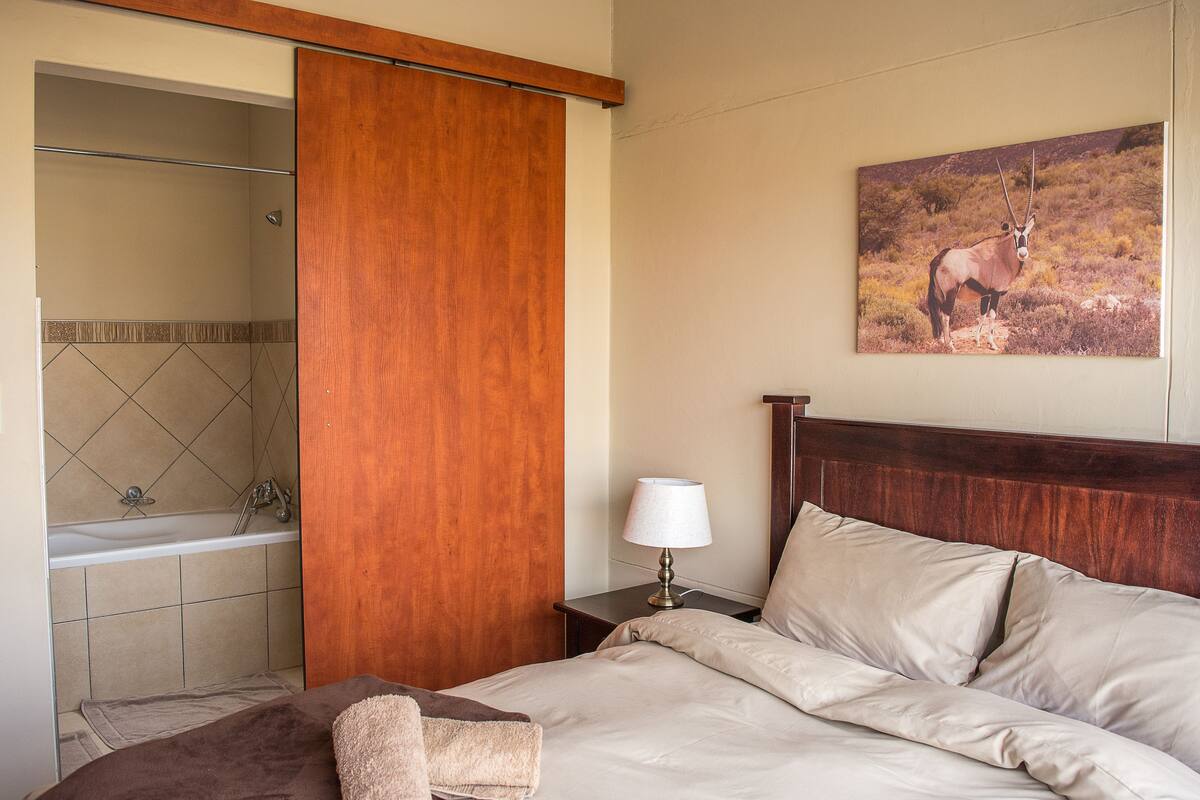 A hotel room with a bed, a nightstand with a lamp, a bathroom with a bathtub, and a wall-mounted picture of an animal.
