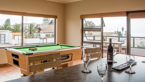 A pool table in a room with large windows, a bar area with glasses and a bottle, and a view of the outside. - Sea View Backpackers - Hostel (Swakopmund)