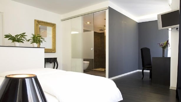 A modern bedroom with a bed, a bedside lamp, a chair, a desk, a mirror, and a sliding door leading to a bathroom. - Pension Mariposa (Weimar)