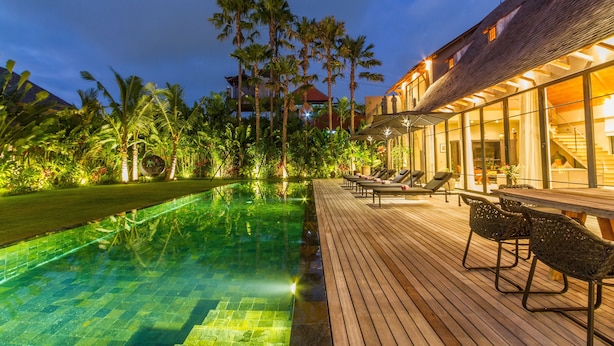 Zona de piscina con cubierta de madera y sillones de descanso, rodeada de abundante vegetación y palmeras.