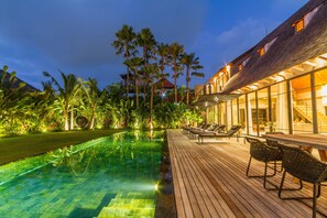 Un espace piscine doté d’une terrasse en bois et de chaises longues, entouré d’une végétation luxuriante et de palmiers.