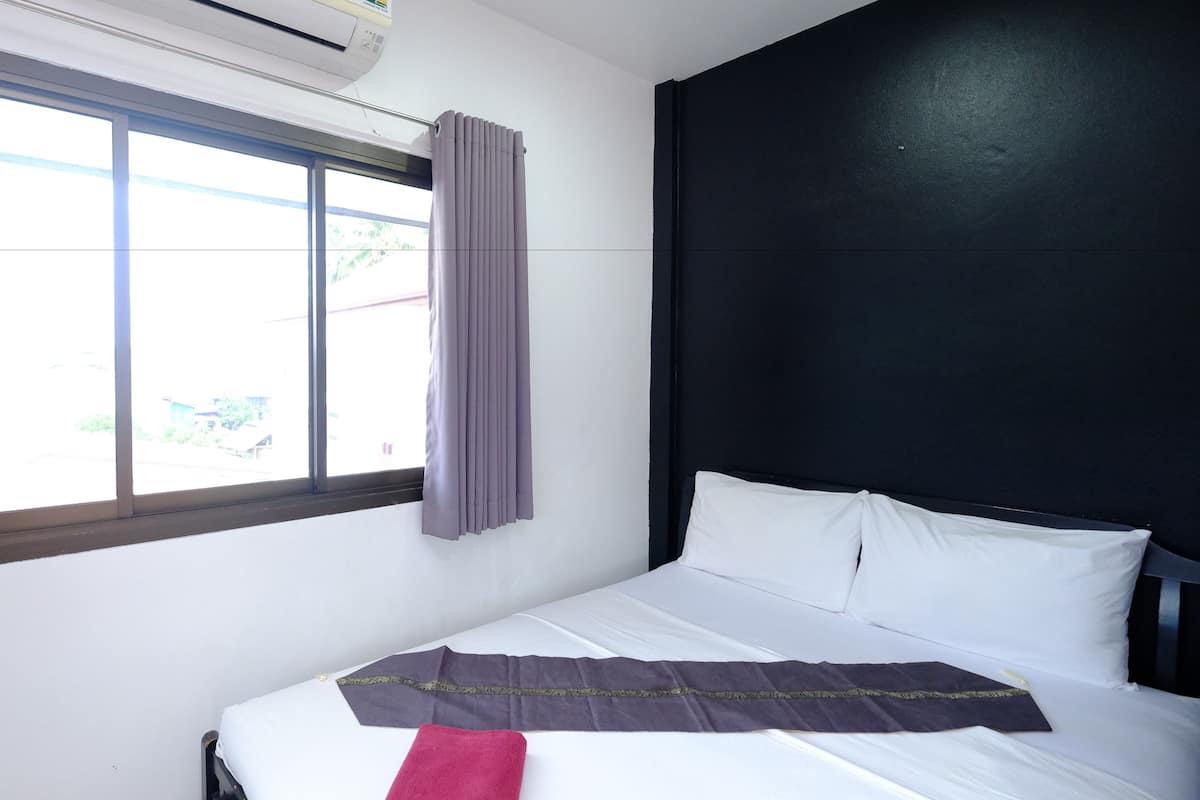 Una habitación de hotel con un ventanal grande, una cama con sábanas blancas, un cabecero oscuro y una manta morada.