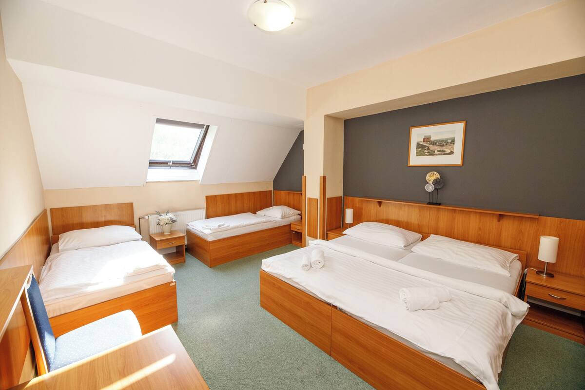 Een hotelkamer met twee eenpersoonsbedden, een tweepersoonsbed, een klein raam, een ingelijst schilderij aan de muur en een nachtkastje met een lamp.