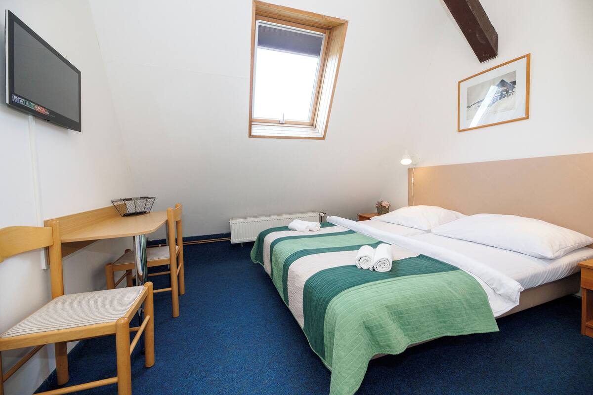 Een hotelkamer met een enkel bed, een bureau met stoel, een klein tafeltje, een aan de muur bevestigde televisie, een raam en een ingelijste foto.