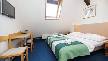 A hotel room with a single bed, a desk with a chair, a small table, a TV mounted on the wall, a window, and a framed picture on the wall.