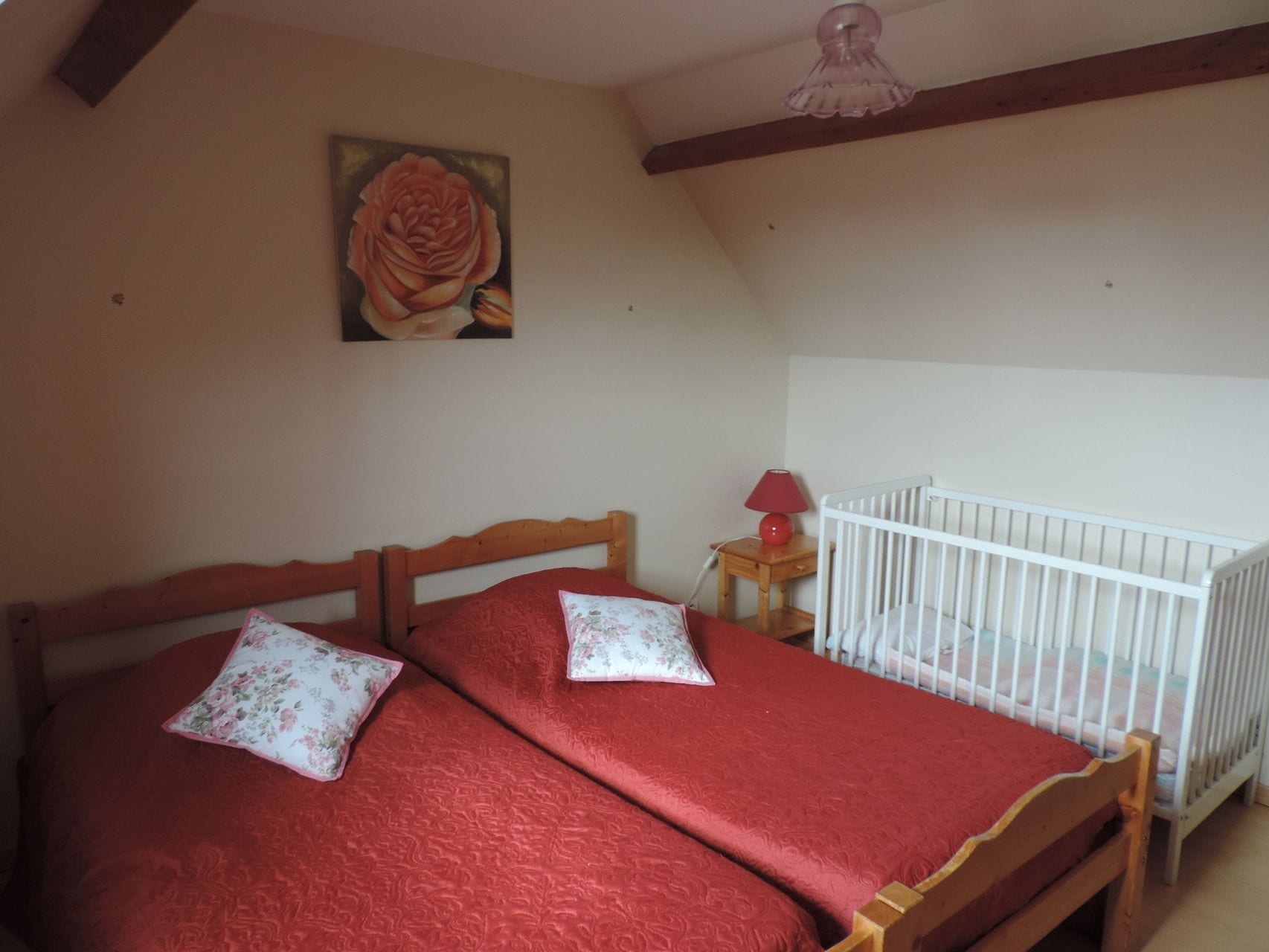 Un dormitorio con una cama de madera, una cuna, una manta roja, un cojín floral y una lámpara roja, además de un cuadro de una rosa en la pared.