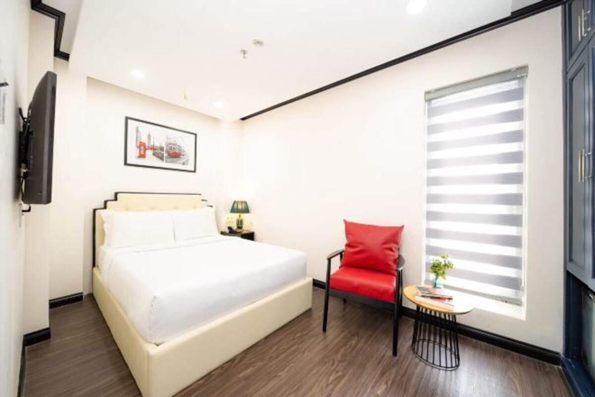 Una habitación de hotel con una cama, una silla roja, una mesita con una planta y una ventana con persianas.